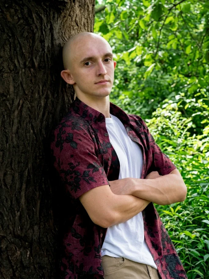 Emmett Dunham, arms crossed, leaning against a tree in summer foliage.