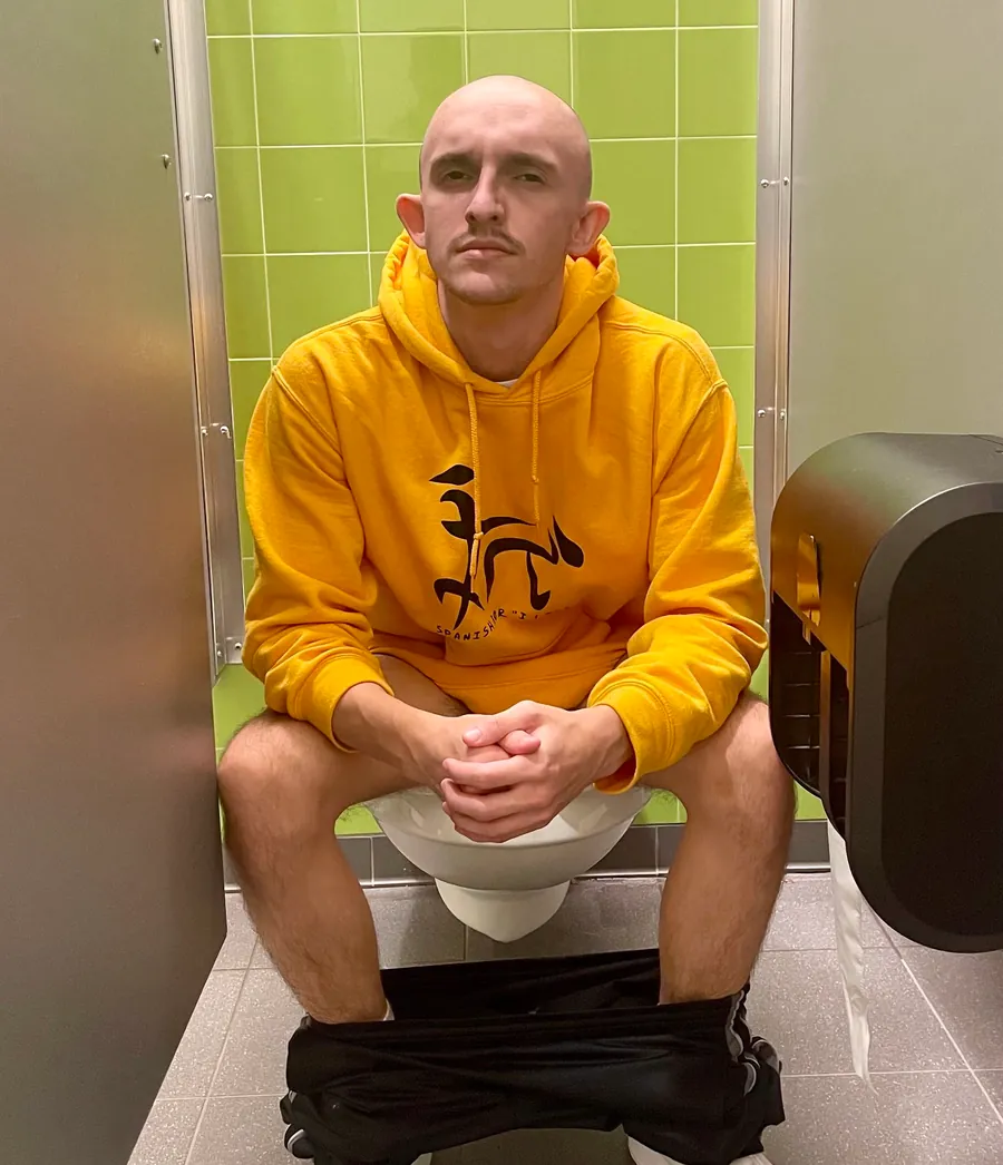 The same figure in the yellow hoodie, seated deadpan in a green-tiled bathroom stall with the lights on.