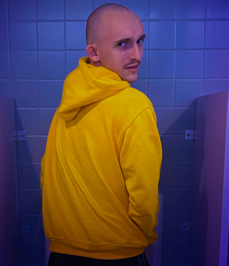 A figure in a yellow hoodie turns to look back, alone between bathroom stalls under blue light.